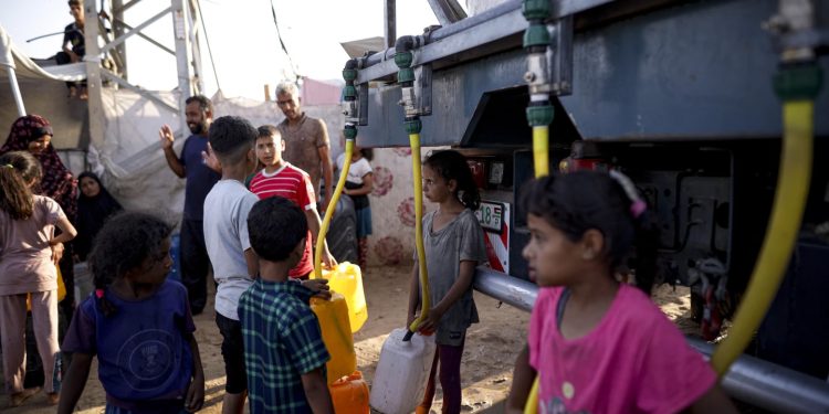 Palestinian children displaced by the Israeli bombardment of the Gaza Strip fill plastic bottles with water as others queue and wait their turn at a makeshift tent camp in the southern town of Khan Younis, Monday, July 1, 2024. (AP Photo/Jehad Alshrafi)ASSOCIATED PRESS